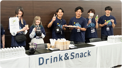 Vue Fes Japan 2024のドリンク＆スナックコーナーで、来場者に飲み物やお菓子を提供するスタッフたち。テーブルの前で笑顔で並んでいる。